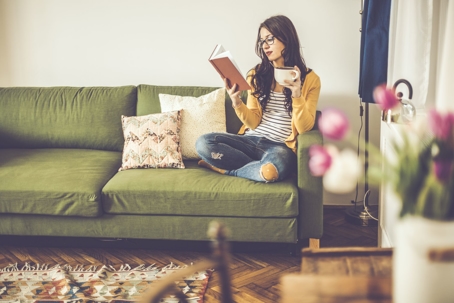 A woman sitting on a couch, reading a book, while sipping coffee